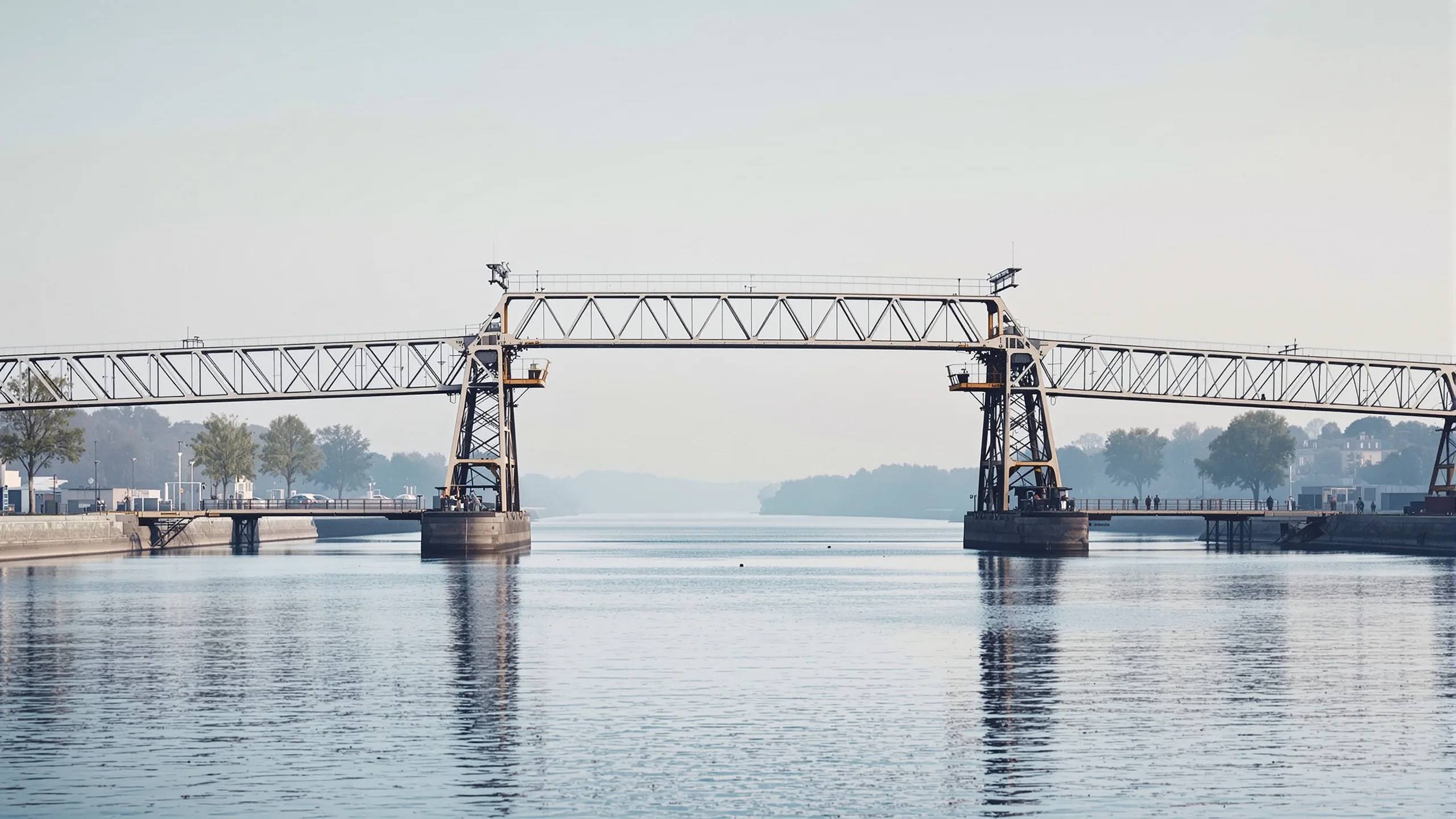 Stählerne Fachwerkbrücke überspannt einen breiten ruhigen Kanal im Morgendunst, Baumreihen an beiden Ufern