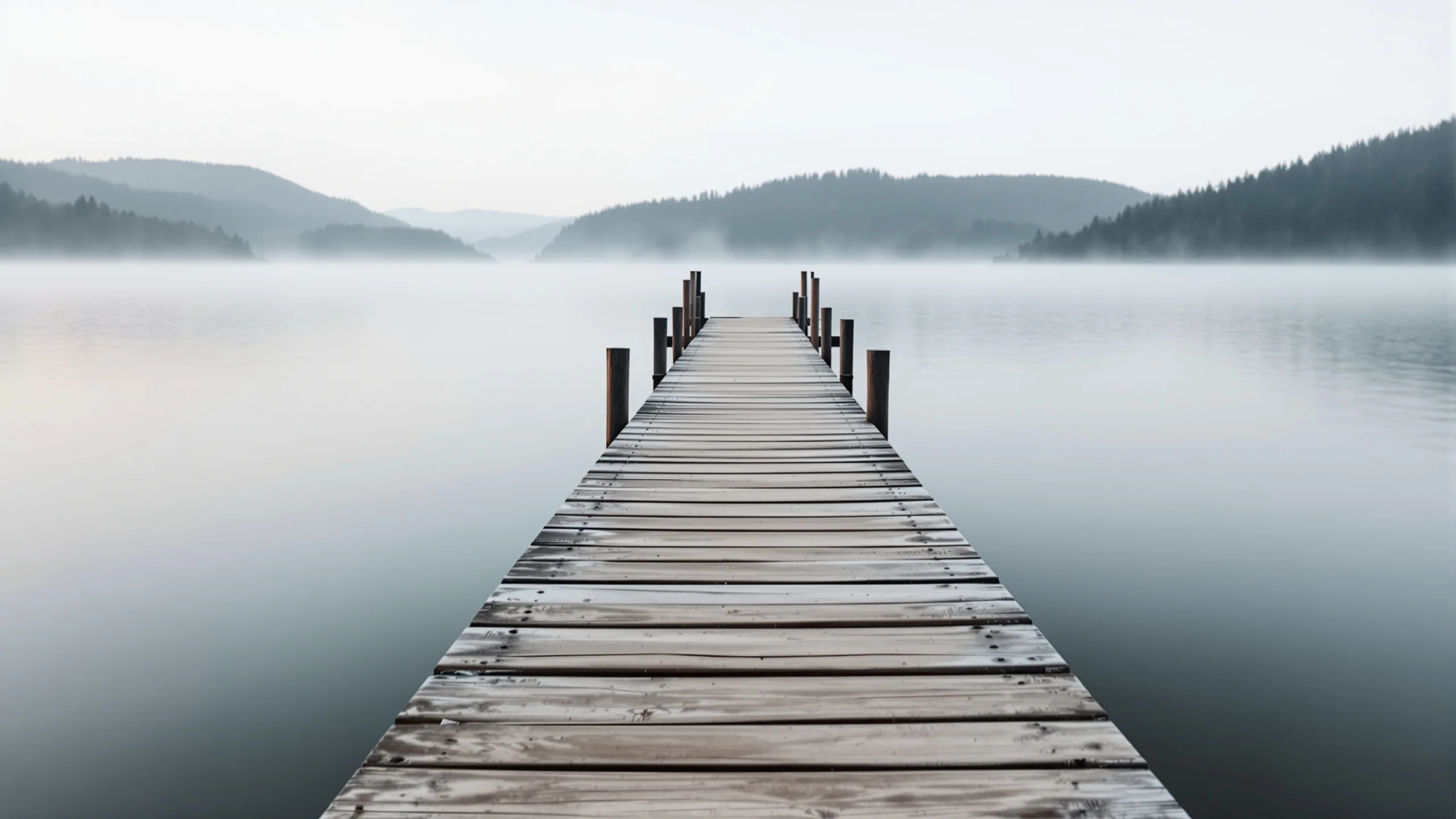 Verwitterter Holzsteg führt in einen völlig stillen See, dünner Nebel liegt auf dem Wasser, bewaldete Hügel am anderen Ufer im ersten Licht